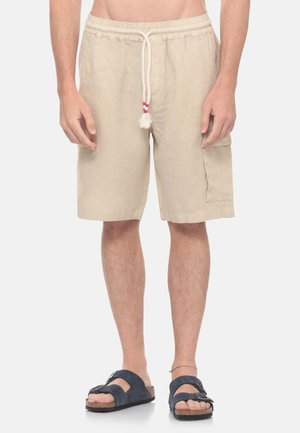 Uomo che indossa pantaloncini beige con coulisse e tasca laterale e sandali blu navy, in piedi su uno sfondo chiaro e semplice.
