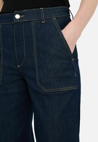 Pantalones de mezclilla azul oscuro con costuras visibles, bolsillos frontales y cierre de botón metálico en la cintura. Cuenta con un ajuste relajado.