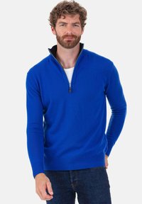 Pull à col zippé bleu en matière douce, avec manches longues et ourlet côtelé. Porté sur un sous-vêtement blanc, avec un col intérieur noir.