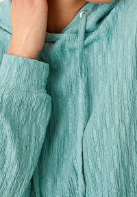 Felpa leggera color turchese con texture, dotata di cappuccio con coulisse e polsini a costine. Il tessuto presenta un motivo ondulato e un design morbido e casual.