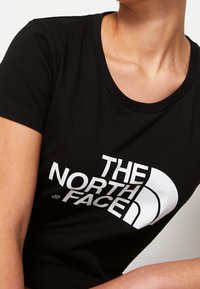 Osoba v černém tričku s krátkým rukávem, na kterém je viditelné bílé logo "THE NORTH FACE" na prsou, viditelném od krku až k horní části paže.