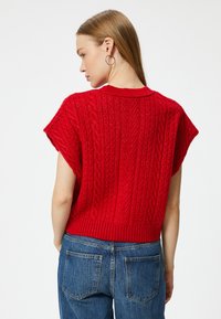 Czerwony sweter dzianinowy z dopasowanym żeberkowym dołem i krótkimi rękawami, z charakterystycznym wzorem w warkocze. Nosi się go z niebieskimi jeansami w wysokim stanie.