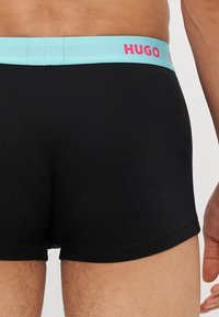 Schwarze Boxershorts mit einem hellblauen Gummibund, der "HUGO" in pinkfarbenen Buchstaben zeigt. Das Material wirkt weich und hat eine glatte Textur.