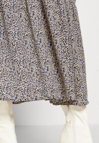 Robe à motif floral avec un fond sombre et des fleurs beiges et bleues. Le tissu tombe doucement ; l'ourlet a un bord froncé. Bottes blanches hautes jusqu'au genou.