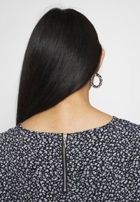 Schwarz-blusige Bluse mit kleinen blauen und weißen Mustern, Rundhalsausschnitt und sichtbar zippbarem Rücken. Das strukturierte Gewebe hebt die Details hervor.