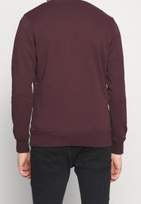 Bordowa bluza z długimi rękawami, ściągaczami na mankietach i u dołu, gładką teksturą oraz okrągłym dekoltem. Noszona z ciemnymi dżinsami.