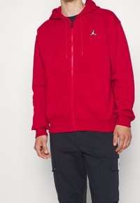 Felpa rossa con zip in misto cotone, dotata di tasca a marsupio e un piccolo logo bianco e nero sul petto. Tessuto morbido e testurizzato.