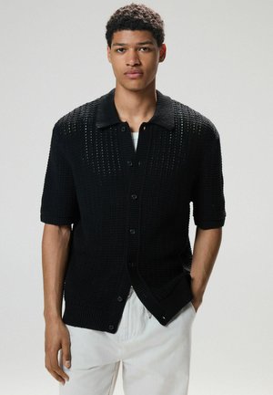 Schwarze gestrickte Kurzarm-Cardigan mit Knopfverschluss, strukturiertem Stoff und Kragen. Kombiniert mit hellen Hosen.