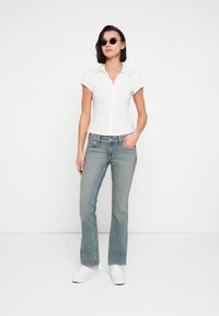 Λευκό στενό πουκάμισο με κοντά μανίκια και κούμπωμα με κουμπιά, συνδυασμένο με ανοιχτό μπλε τζιν με καμπάνα. Το μοντέλο φοράει μαύρα γυαλιά ηλίου και λευκά αθλητικά παπούτσια.