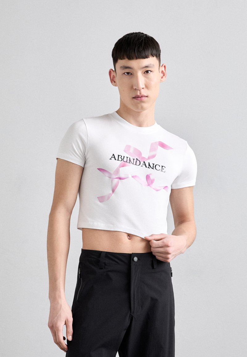 T-shirt blanc court à manches courtes, avec le mot "ABONDANCE" en texte noir et des graphiques de rubans roses sur le devant. Pantalons noirs avec poches.