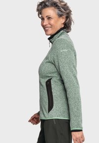 Grüner melierter Fleece-Jacke mit hohem Kragen, Reißverschluss-Brusttasche und seitlichen Taschen sowie schwarzen Akzenten. Ausgestattet mit langen Ärmeln und enganliegenden Bündchen.