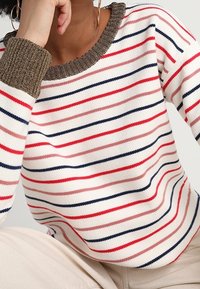 Pull-over en maille avec un col rond, présentant des rayures horizontales en rouge, marine et rose clair sur un fond crème. Poignets côtelés marron.