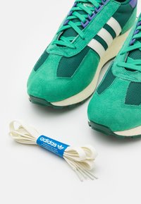 Gröna mocka- och mesh-idrottsskor med vita r änder, med lila detaljer och en texturerad sula. Inkluderar vita snörningar med blå logotyp.