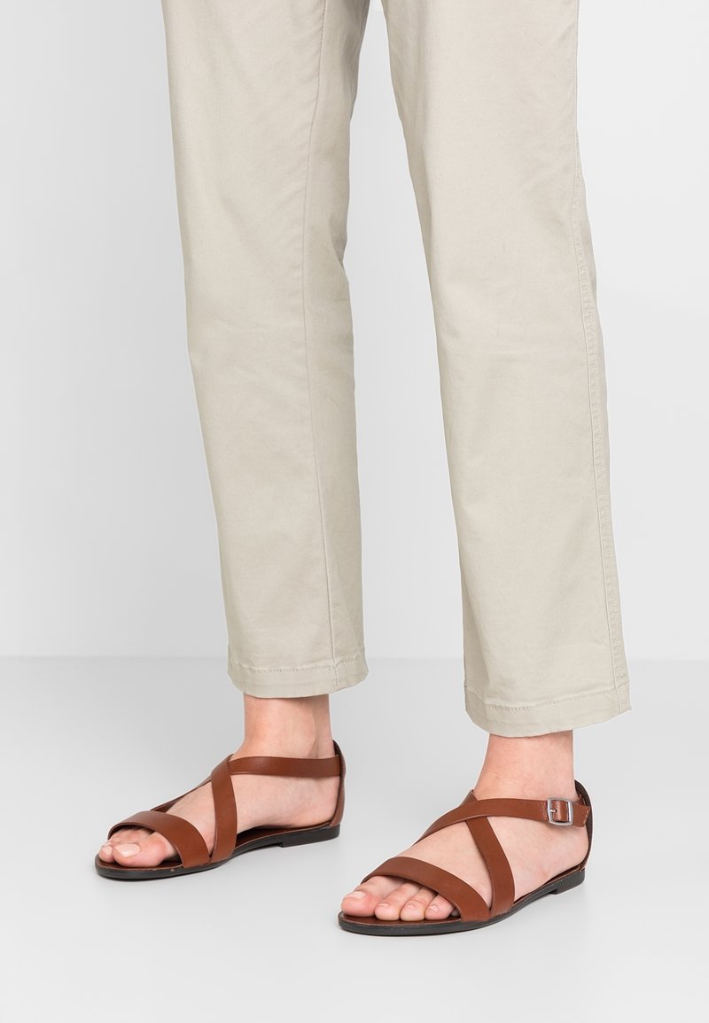 Sandales en cuir marron avec design à lanières croisées et boucle à la cheville, associées à un pantalon court beige clair. Texture lisse et style minimaliste.