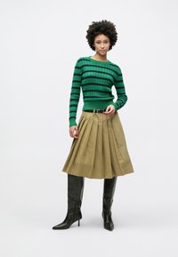 Πλεκτό πουλόβερ με πράσινες και μαύρες ρίγες, μπεζ πλισέ φούστα και μαύρες δερμάτινες μπότες μέχρι το γόνατο. Απλή ζώνη τονίζει τη μέση.