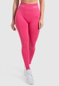Leggings rosa con un alto cinturino che mostrano il testo "SMIL". Il tessuto è liscio con un design aderente e elastico, perfetto per l'abbigliamento sportivo.