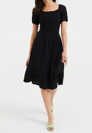 Mujer con un vestido midi negro de ojales, con mangas abullonadas y escote cuadrado, combinado con tacones abiertos en color verde claro.