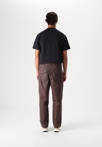 T-shirt nera in cotone e pantaloni marroni in cotone con tasche posteriori, dotati di polsini regolabili. L'outfit è completato da sneakers bianche.