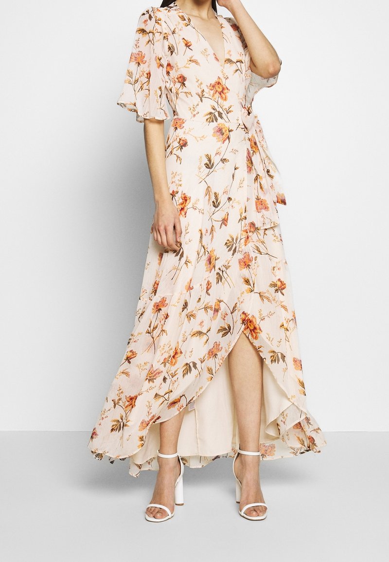 Robe maxi florale en rose pastel avec des manches courtes volantées, un design croisé et un ourlet asymétrique. Des sandales à blocs blanches complètent le look.
