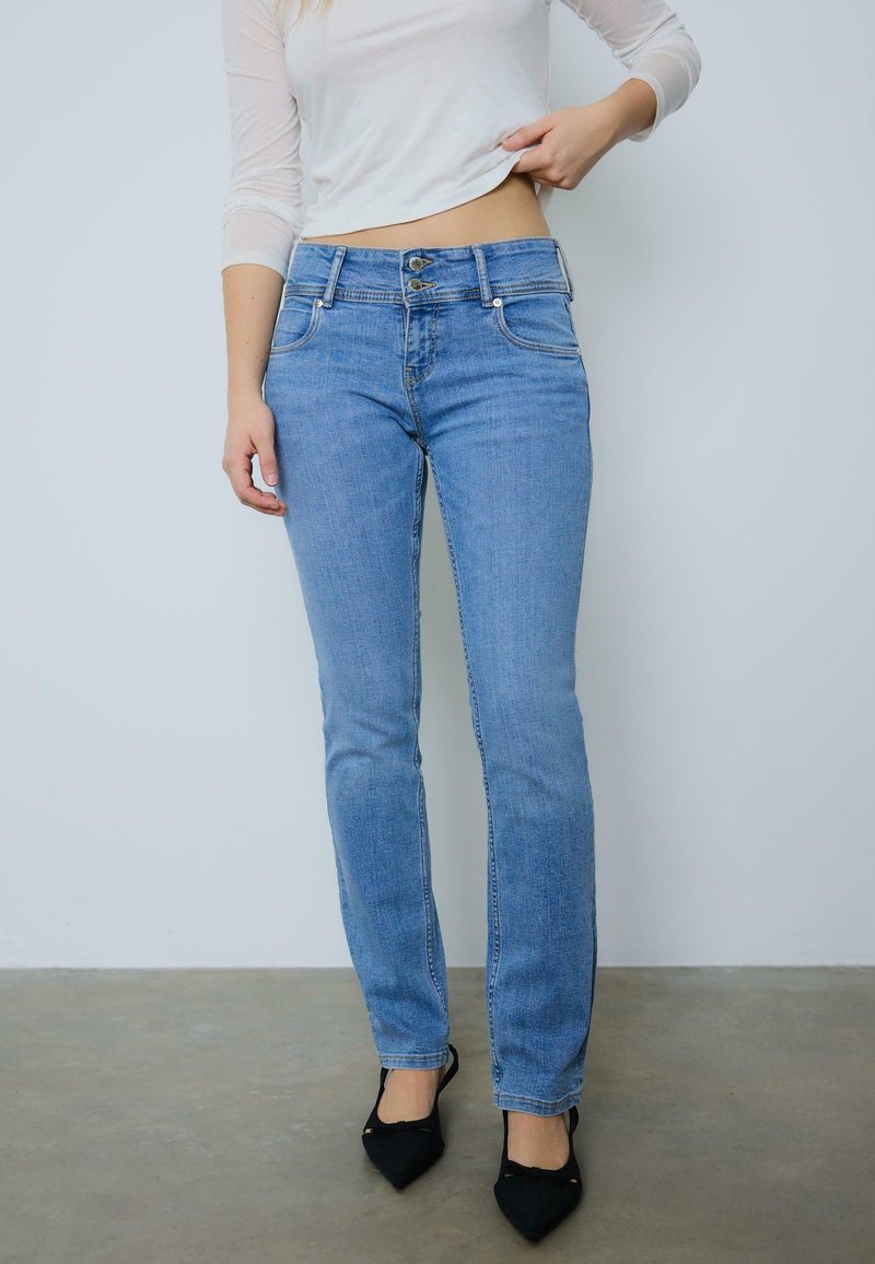 Personne portant un jean bleu clair à double boutonnage, un haut blanc à manches longues partiellement relevé, et des chaussures noires à bout pointu, debout sur un sol gris.