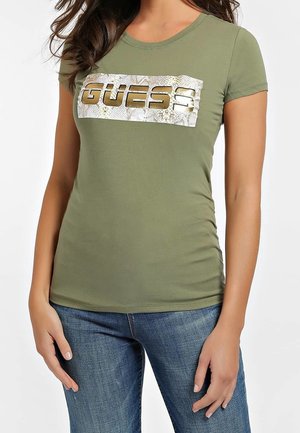 Vrouw draagt een olijfgroene korte mouwen shirt met een metalen "GUESS" logo en blauwe jeans, staat met ontspannen handen langs haar zijde.