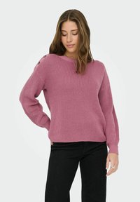 Jeune femme portant un pull en maille mauve avec des détails de boutons sur l'épaule et un pantalon noir, debout avec une main sur la hanche.