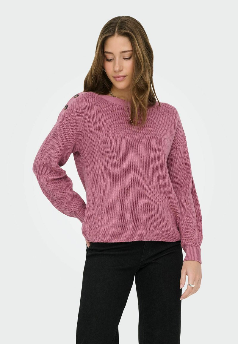 Jeune femme portant un pull en maille mauve avec des détails de boutons sur l'épaule et un pantalon noir, debout avec une main sur la hanche.