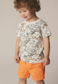 Maglietta bianca con stampe di disegni colorati, tra cui sole e animali, abbinata a pantaloni corti arancioni. Realizzata in cotone morbido per un comfort ottimale.