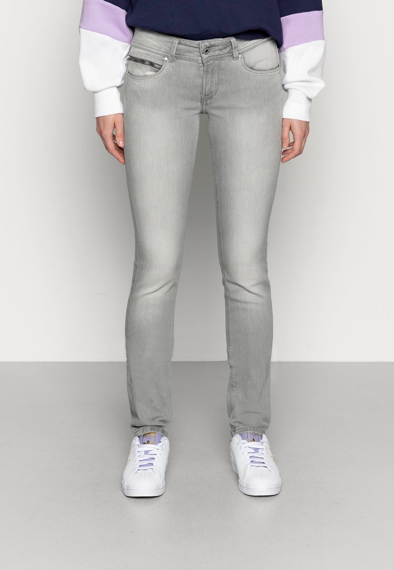 Šedé skinny džíny s hladkou texturou, tradičním designem pěti kapes a decentním vyblednutím; spárovány s bílými teniskami s fialovými akcenty.