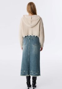 Haut en tricot beige coupé avec une capuche et un ourlet festonné, associé à une longue jupe en denim bleue avec des bords effilochés et un léger aspect vieilli.