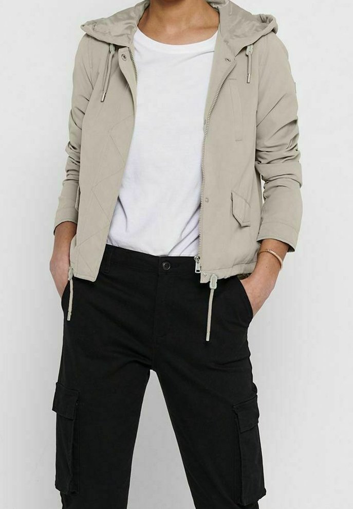 Personne portant une veste beige zippée à capuche sur une chemise blanche et un pantalon cargo noir, mains dans les poches.