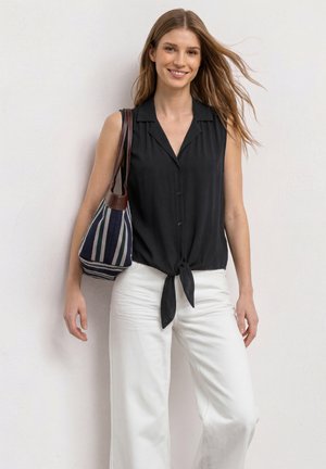 Jeune femme aux cheveux châtain clair porte un chemisier noir sans manches noué devant, un pantalon blanc et porte un sac cabas en tissu rayé sur l'épaule.