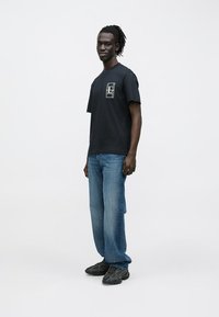 Μαύρο T-shirt με μικρό γραφικό στο αριστερό στήθος, συνδυασμένο με ανοιχτό μπλε τζιν και μαύρα αθλητικά παπούτσια. Το μοντέλο στέκεται όρθιο μπροστά σε απλό φόντο.