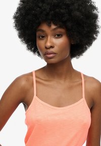 Camisole color pesca con spalline sottili, texture morbida, leggero luccichio e scollo arrotondato, evidenziata su uno sfondo neutro.