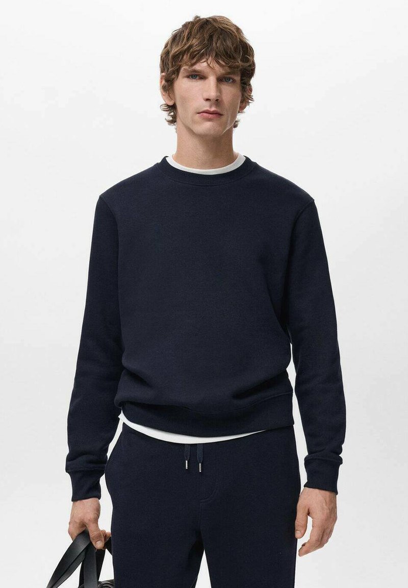 Marineblauwe crewneck sweatshirt van een katoenmix, met geribbelde manchetten en zoom. Draagt over een wit overhemd, gecombineerd met bijpassende broek en een zwarte tas.