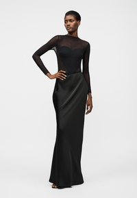 Femme portant une longue robe noire avec un haut en maille transparente et une jupe unie, debout avec une main sur la hanche devant un fond clair uni.