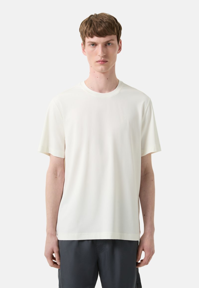 Jonge man in een eenvoudige witte T-shirt met korte mouwen en een ronde hals, en donkere, relaxte broek, staande tegen een lichtgrijze achtergrond.