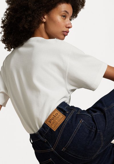 Vit bomulls-t-shirt med korta ärmar, i kombination med mörkblå jeans i denim med en tan läderpatch som har "Polo" broderat på midjebandet.