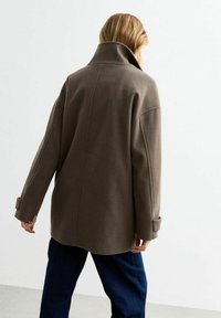 Cappotto oversized in lana marrone con colletto alto, spalle scese e polsini abbottonati, caratterizzato da una texture liscia e cuciture minime.