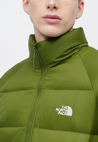 Zaļa polsterēta jaka ar augstu apkakli, priekšpusē rāvējslēdzējs un balts logotips kreisajā krūtīs. Gluda tekstūra un viegls dizains.