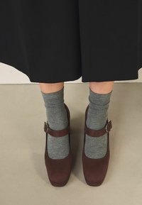 Pantofi din suede maro în stil Mary Jane, cu vârful rotunjit și un detaliu mic de cataramă, purtați cu șosete din lână gri, așezați pe un fundal neutru.