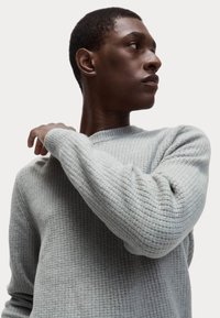Suéter gris claro en un tejido texturizado y acanalado con cuello redondo y mangas largas. La modelo posa con la mano en el hombro.