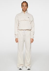 Beige träningsoverall i slätt tyg; har en dragkedjekrage, elastisk midja och sidostrand. Kombinerad med vita sneakers.