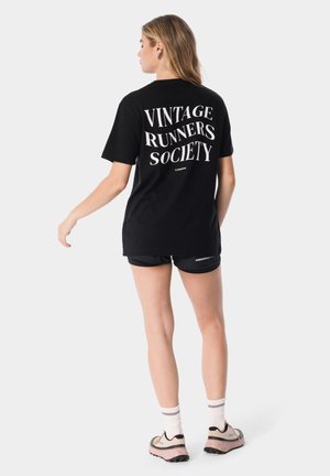 Kobieta ubrana w czarną koszulkę "Vintage Runners Society", czarne spodenki, białe skarpetki i różowe buty do biegania stoi tyłem zwrócona do przodu.