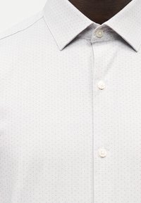 Λευκό πουκάμισο με μικρό πουά σχέδιο, κουμπωτό γιακά και δύο ορατά λευκά κουμπιά στην εμπρόσθια πατιλέτα.