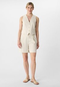 Cream-colored, sleeveless vest with a V-neck and three buttons, paired with matching shorts. Simple design and textured fabric. Worn with woven slide sandals.