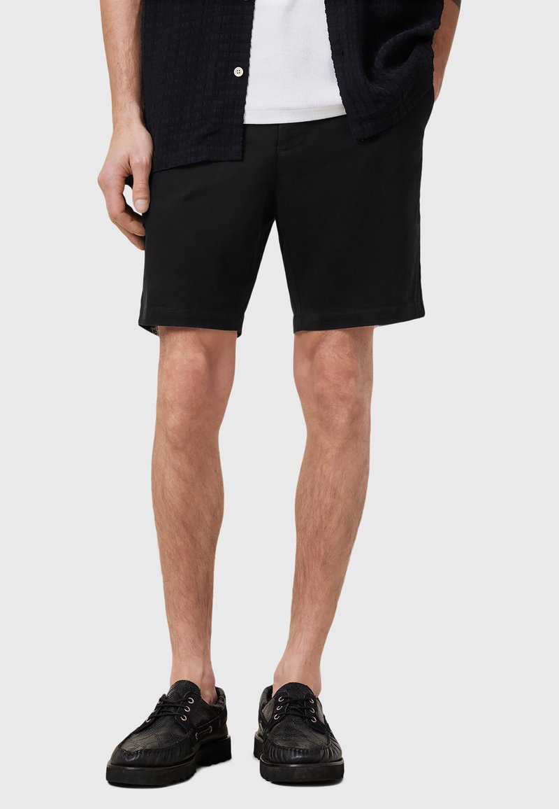 Pantaloni shorts neri con una texture liscia, di lunghezza sopra il ginocchio, abbinati a scarpe nere con lacci e un design a motivo.
