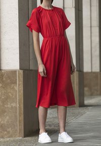 Robe rouge en tissu léger, avec des manches courtes, un col boutonné et une taille élastique. Associée à des baskets blanches.