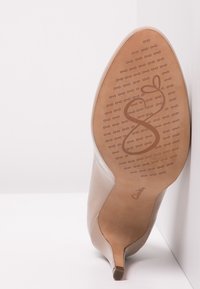 Chaussure à talon haut beige avec un bout pointu, tige en cuir verni lisse et semelle en caoutchouc marron ornée d'un motif décoratif et d'un logo.