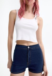 Mujer con cabello rosa que lleva una camiseta blanca corta con adornos en forma de bola y pantalones cortos de mezclilla oscuros de tiro alto.
